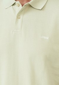 Lichtgroene polo gemaakt van gestructureerde stof, met een opgestikte kraag, twee knopen en een klein wit logo aan de linkerkant.