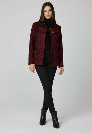 Abrigo texturizado de cuadros burdeos con dos bolsillos delanteros y grandes botones, combinado con un jersey de cuello alto negro y vaqueros ajustados, completado con botines de tobillo.