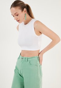Witte cropped tanktop gecombineerd met lichtgroene high-waisted jeans. De tanktop heeft een gladde textuur en een ronde halslijn.