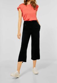 Korálová blůza s krátkým rukávem a volány, spárovaná s černými kalhotami s širokými nohavicemi a bočními kapsami, a bílými platformovými teniskami.