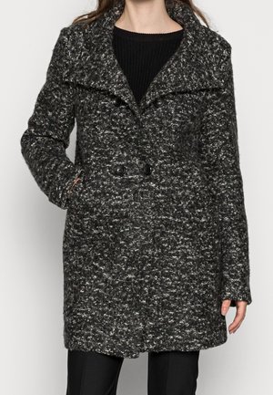 Woman wearing a black and gray textured double-breasted winter coat with wide lapels and side pockets, over a black sweater.