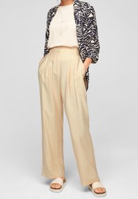 Marinblå och krämfärgad mönstrad blazer över en krämfärgad topp; plisserade beige byxor och ljusa sandaler med korksål. Flytande former och mjuka texturer.