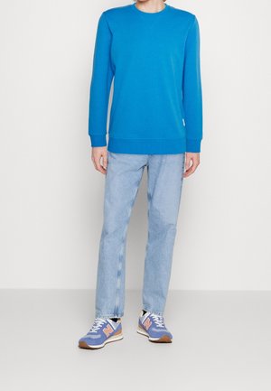 Felpa blu con maniche lunghe e polsini a coste, abbinata a jeans azzurri e sneakers blu New Balance con dettagli bianchi.