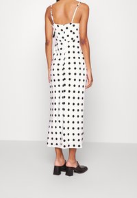 Kvinna klädd i en vit ärmlös midikjol med svarta prickar och svarta klacklösa skor med blockklackar, står vänd med ryggen mot kameran.