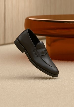 Mocassino in pelle nera con tacco basso, dettagli cuciti e cinturino con logo, posizionato su tappeto beige vicino a un tavolo in legno rotondo.