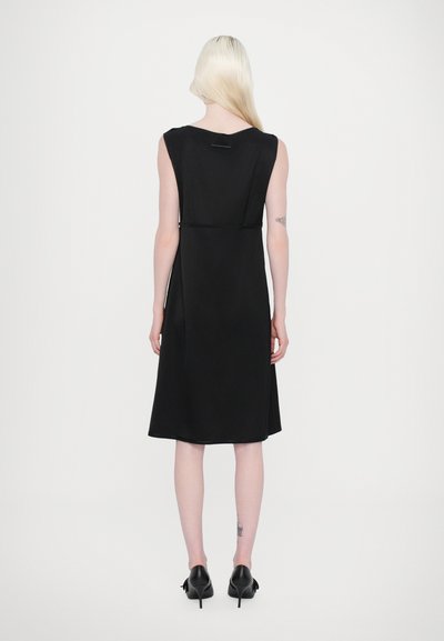 Robe noire sans manches avec une texture lisse, longueur genoux, et une découpe subtile au dos, assortie à des chaussures à talons noires.