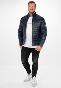 Marine blauwe gewatteerde jas met een hexagonaal patroon, opstaande kraag, ritssluiting en zijzakken, gedragen over een witte shirt en zwarte jeans.