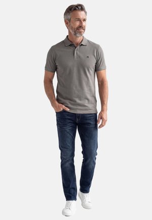 Mann mit grauen Haaren und Bart, trägt ein graues Polohemd, dunkelblaue Jeans und weiße Turnschuhe, steht mit einer Hand in der Tasche.