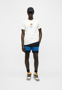 Νεαρός άνδρας που φοράει μαύρο καπέλο Nike, λευκό μπλουζάκι με γραφικό κούπα καφέ, μπλε και μαύρη σορτς, μαύρες κάλτσες και πολύχρωμα αθλητικά παπούτσια.