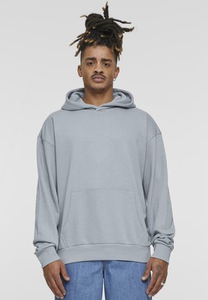 Lys grå hettegenser laget av myk stoff; har en frontlomme, justerbar hette og ribbestrikkede mansjetter. Båret med blå denimjeans.