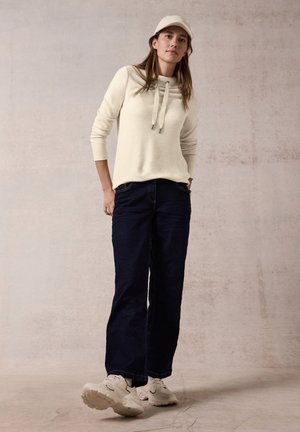 Un pull crème avec un col roulé et des cordons, associé à un jean large navy et des baskets blanches, sur un fond neutre.