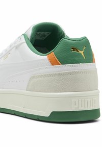 Zapatilla blanca con acentos verdes, talón texturizado verde, detalle naranja y logo dorado. Suela blanca gruesa con ribete verde.