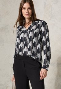 Blusa patterned nera e grigia con scollatura a V, maniche lunghe e tessuto morbido, abbinata a pantaloni neri. Design minimalista e moderno.