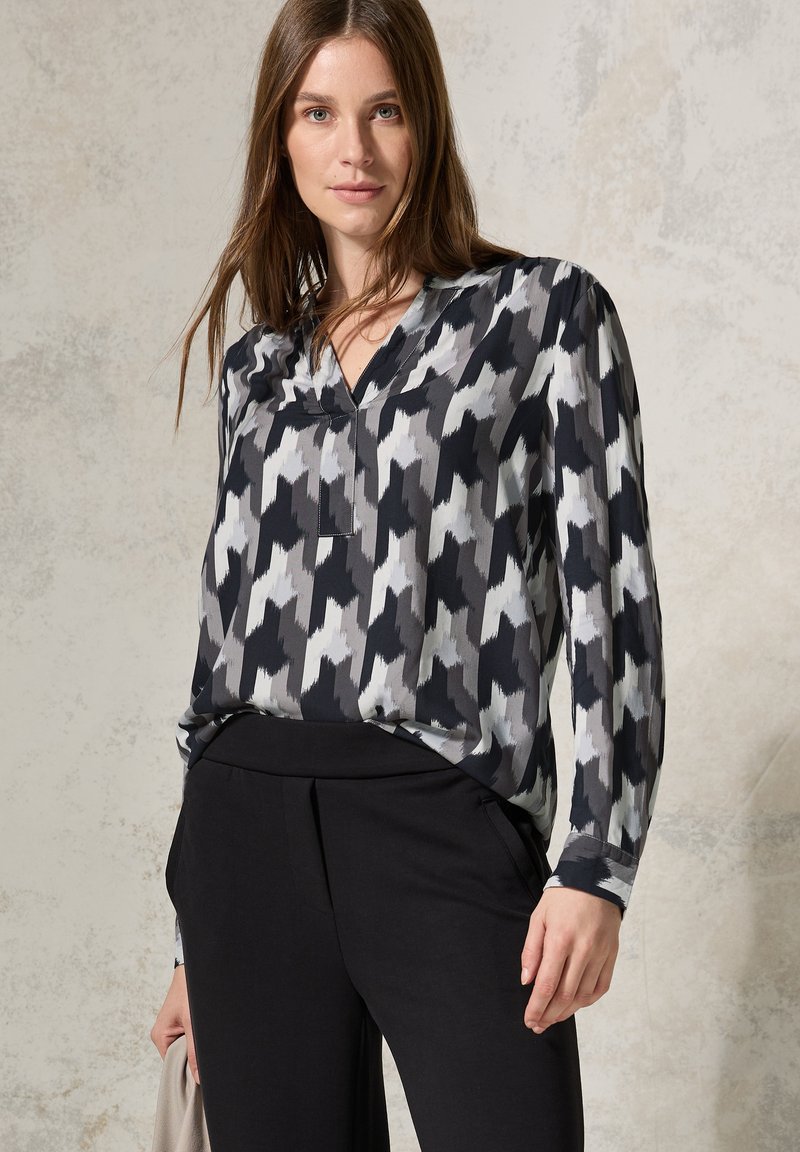 Blusa patterned nera e grigia con scollatura a V, maniche lunghe e tessuto morbido, abbinata a pantaloni neri. Design minimalista e moderno.
