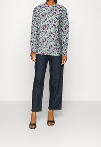 Pitkähihaispaita, jossa on violetteja ja vihreitä kukkakuoseja, yhdistettynä tummansinisistä farkkukankaasta tehtyihin suoralahkeisiin housuihin ja mustiin avokkaisiin avoimilla varpailla.