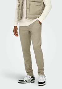 Pantaloni della tuta beige con tasche laterali, abbinati a un maglione leggero e a un gilet imbottito. Completati da scarpe da ginnastica nere e bianche.