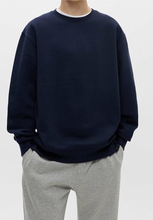 Personne portant un sweat-shirt bleu marine uni et un pantalon de survêtement gris clair, debout les mains dans les poches devant un fond uni.