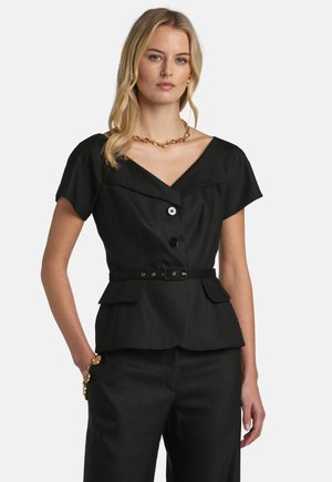 Kvinde iført en sort kortærmet blazer med bælte og to sorte knapper samt matchende sorte bukser mod en ensfarvet baggrund.