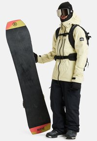 Snowboard mit einer schwarzen Oberfläche und rot-gelben Akzenten, gehalten von einer Person in einer hellgelben Jacke und schwarzen Hosen. Schwarze Handschuhe und eine Ski-Brille.