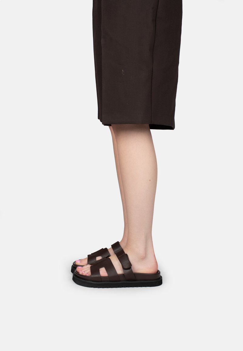 Piernas con falda negra hasta la rodilla y sandalias marrón oscuro de dos tiras con hebillas ajustables en suelas planas.