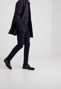 Cappotto lungo blu navy sopra un maglione scuro, abbinato a pantaloni cargo scuri e scarpe nere con lacci. Texture lisce e dettagli sottili visibili.