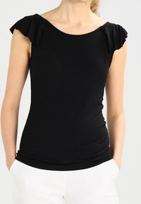 Žena v přiléhavém černém blůzce s krátkým rukávem a nabíranými rameny, v bílých kalhotách, s rukama v kapsách, na jednobarevném pozadí.