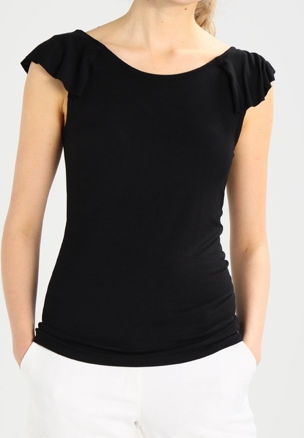 Žena v přiléhavém černém blůzce s krátkým rukávem a nabíranými rameny, v bílých kalhotách, s rukama v kapsách, na jednobarevném pozadí.