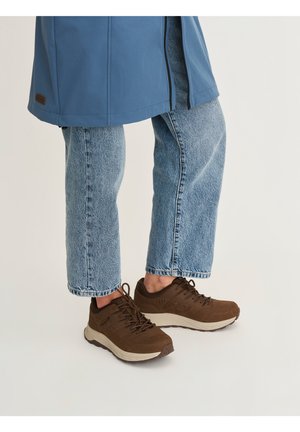 Persoon draagt lichtblauwe spijkerbroek, bruine sneakers met witte zolen en een blauwe jas, staand tegen een effen lichte achtergrond.