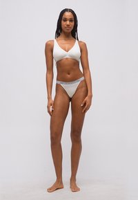 Γυναίκα όρθια, ξυπόλητη, φοράει λευκό σουτιέν και λευκά εσώρουχα με τη μάρκα Emporio Armani, μπροστά σε απλό γκρι φόντο.