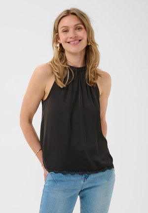 Femme souriante aux cheveux blonds ondulés portant un haut noir sans manches avec bordure en dentelle et un jean bleu clair sur un fond uni.