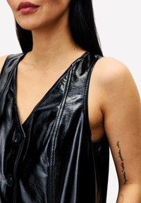 Frau mit langen dunklen Haaren, die ein ärmelloses schwarzes Lederoberteil trägt und ein Tattoo mit senkrechem Schriftzug am Oberarm zeigt.