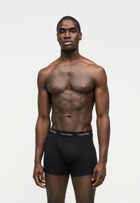 Αθλητικός άνδρας με σκουρόχρωμο δέρμα, στέκεται μπροστά σε απλό φόντο, φοράει μαύρα boxer Calvin Klein, κοιτάζοντας μπροστά με ουδέτερη έκφραση.