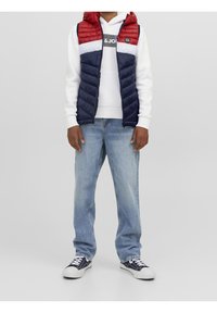 Person som bär en röd, vit och marinblå puffermantel över en vit hoodie, ljusblå jeans och svarta och vita sneakrar, står framför en grå bakgrund.