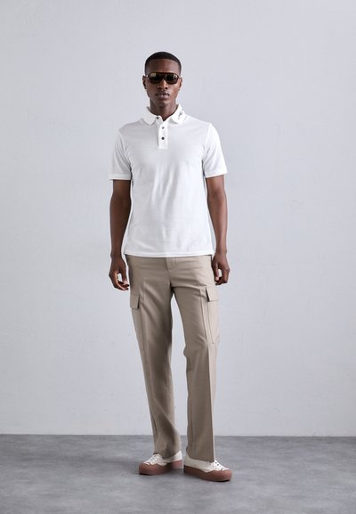 Hvit polo skjorte med tre-knapps krage, beige cargo-bukser med sidelommer, og hvite sneakers med rosa såle. Solbriller på.