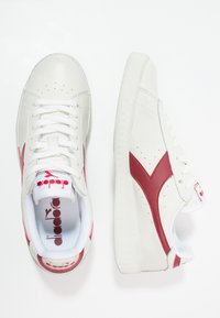Sneakers bianchi con tomaia in pelle texture, dettagli rossi e logo sulla linguetta. Presentano una punta perforata e una suola in gomma spessa.