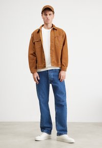 Bruine denim jas over een witte sweater, gecombineerd met blauwe jeans en witte sneakers. De outfit is compleet met een bijpassende bruine pet.