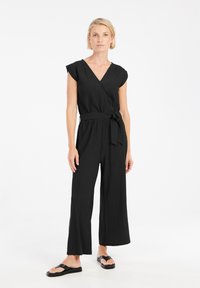 Zwarte wijde jumpsuit met een V-hals en korte mouwen, voorzien van een ceintuur in de taille en een gestructureerde stof. Gecoördineerd met eenvoudige zwarte sandalen.