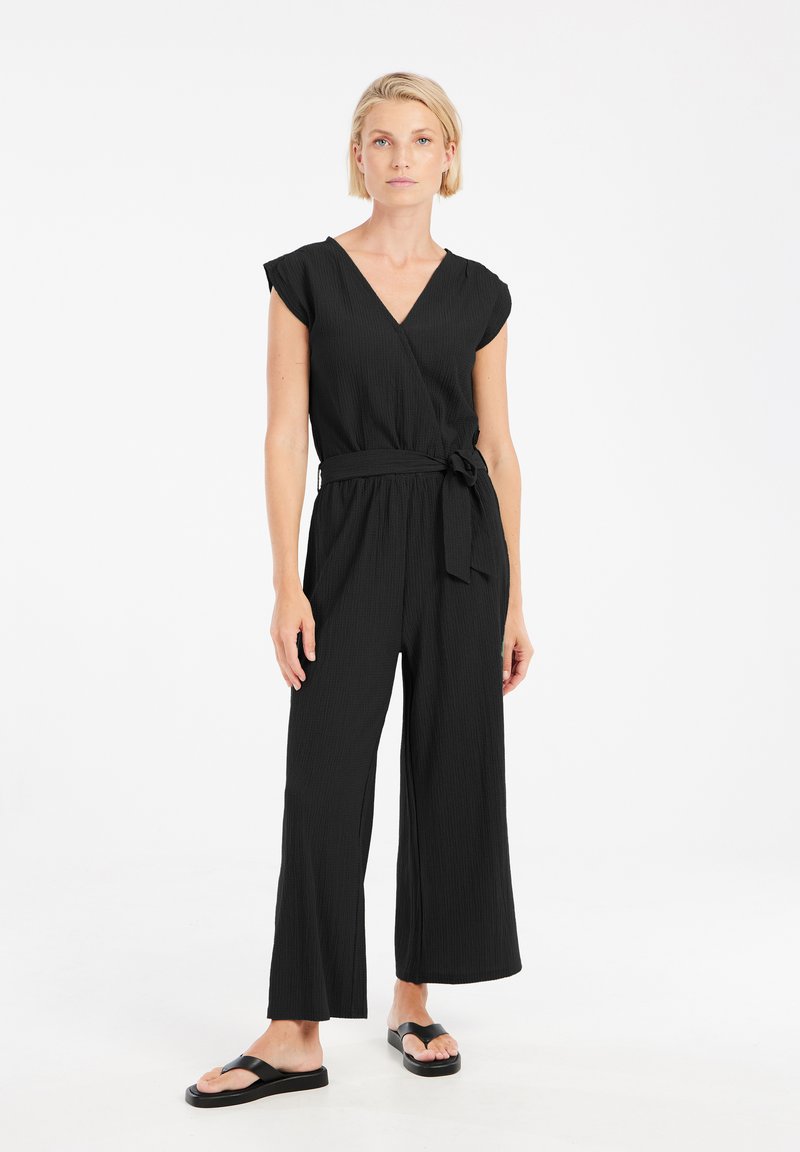 Zwarte wijde jumpsuit met een V-hals en korte mouwen, voorzien van een ceintuur in de taille en een gestructureerde stof. Gecoördineerd met eenvoudige zwarte sandalen.