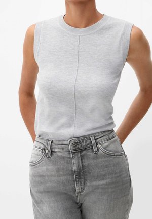 Kvinde iført en ærmeløs lysegrå strikbluse stoppet ned i højbenede falmede grå jeans, stående med hænderne bag ryggen.