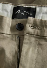 Pantalones caqui con cierre de cremallera y botón. La tela es texturizada con un forro interno suave y una etiqueta negra con el nombre de la marca.
