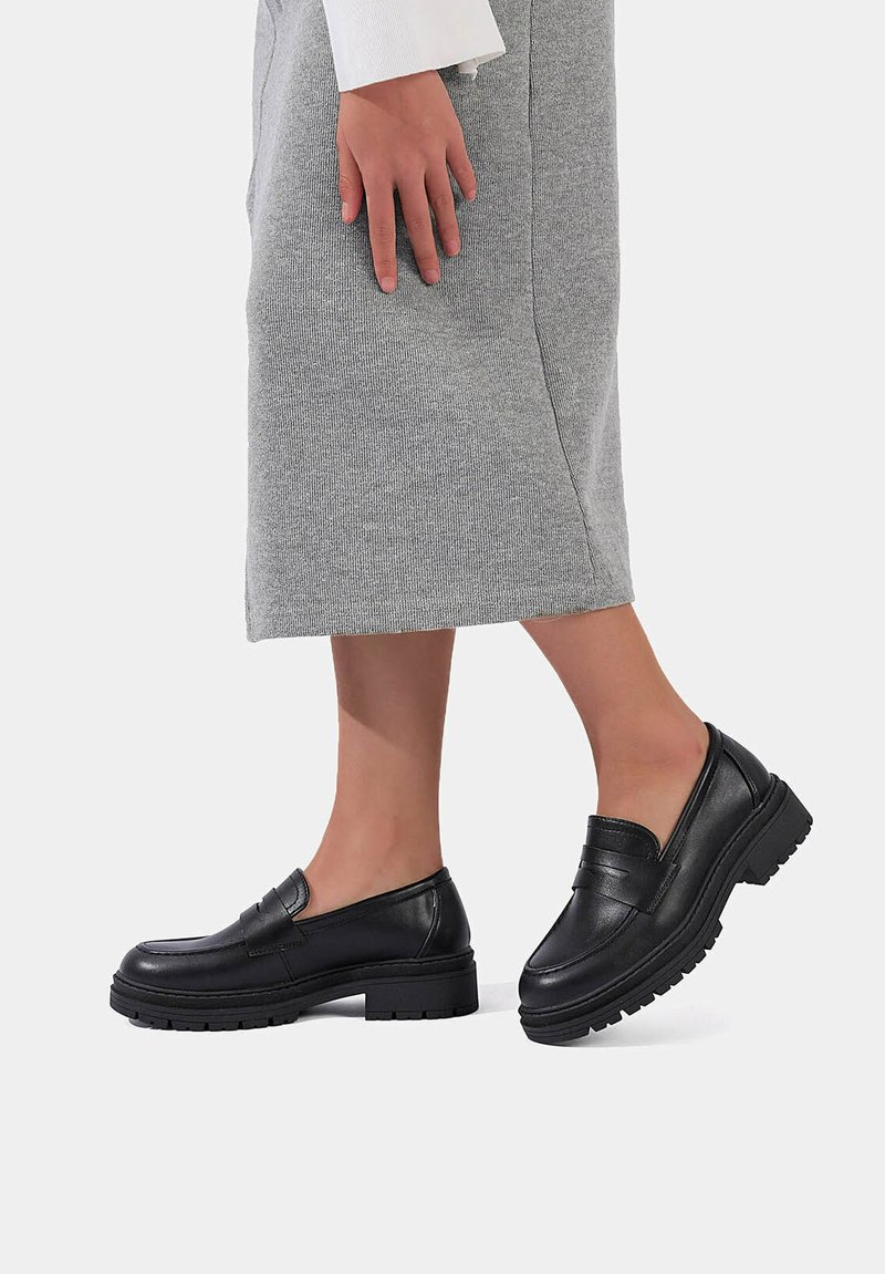 Mocassini in pelle nera con suola spessa, caratterizzati da una finitura liscia e una punta tonda. Indossati con una gonna grigio chiaro.