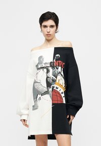 Γυναίκα με μαύρο και λευκό φόρεμα σε υπερμεγέθη γραμμή με ανοιχτούς ώμους, στο κέντρο του οποίου υπάρχει γραφικό με παίκτη μπάσκετ και διαχωρισμένο κείμενο.