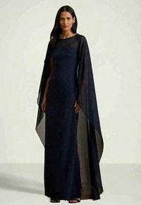 Nő egy hosszú, sötétkék estélyi ruhában, áttetsző, lebegő köpenyujjúval, egy világos háttér előtt.