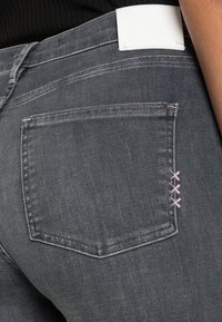 Šedé džíny z denimu mají zadní kapsu s růžovým prošíváním a bílou nášivku na pase. Tkanina má jemnou strukturu.