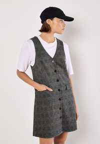Vestido a rayas grises con diseño de escote en V, botones frontales y bolsillos laterales. Lleva una camiseta blanca debajo y combina con una gorra negra.