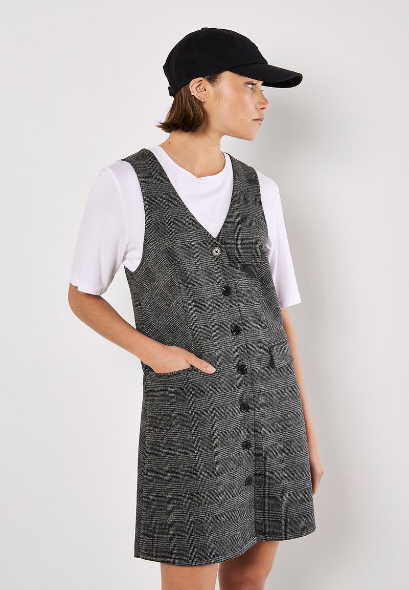 Vestido a rayas grises con diseño de escote en V, botones frontales y bolsillos laterales. Lleva una camiseta blanca debajo y combina con una gorra negra.