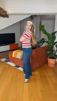 Fiatal nő csíkos ingben és farmerban forog a fapadlón egy kényelmes nappaliban, ahol narancssárga kanapé és nagy cserepes növény található.