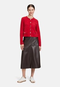 Kobieta ubrana w czerwoną, zapiętą na guziki, puszystą kardigan, ciemnobrązową skórzaną spódnicę midi i białe trampki, stojąca na jednolitym tle.