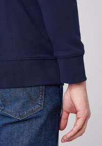 Marinblå sweatshirt med ribbad mudd, ihop med blå jeans som har en utmärkande V-söm i fickdesign. Texturerad tygdetalj synlig.