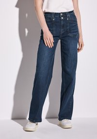 Jeans de tiro alto en azul oscuro con un diseño de pierna ancha, que cuentan con bolsillos frontales y un cierre de botones, combinados con zapatillas blancas.